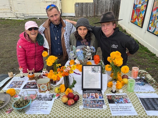 Arinna stands with a group of 3 friends behind an alter created in Fort Bragg, CA to raise awareness of the genocide in Gaza. The table is adorned with flowers and various images of residents of Gaza. In the background, artwork is visible on the wall of a neighboring building.