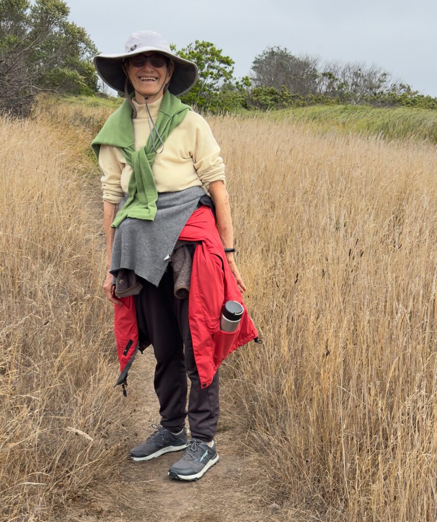 A photo of Arinna walking on a path through dried grass, near the ocean, which is out of sight. She's wearing hiking garb including a hate and sunglasses. She's got a big smile on her face.