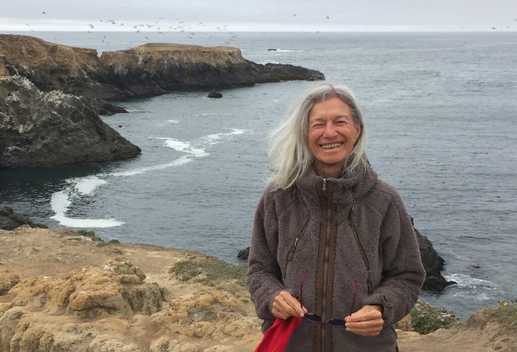 A photo of Arinna Weisman smiling radiantly on a cliff overlooking the Pacific ocean. The seas are calm and gulls flock in the distance. Arinna's wearing a grey fleece jacket and holding her sunglasses and a hat at her waist.